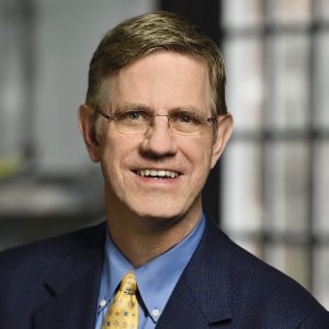 A headshot of Kevin Hemker wearing an indigo suit with a blue shirt and a tie.