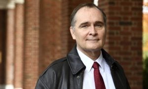 A headshot of James Bellingham wearing a leather jacket and standing in front of brick pillars in the background